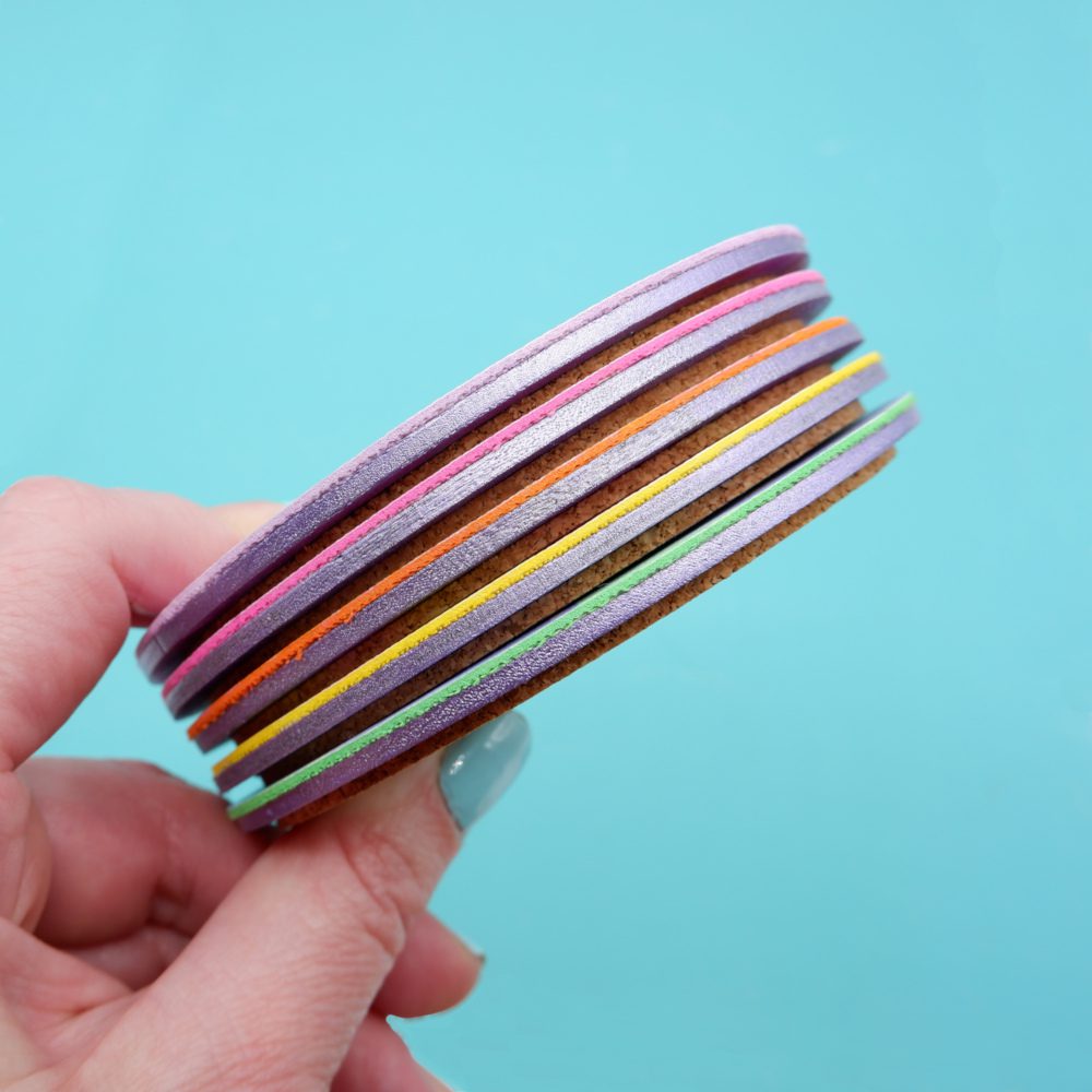A set of round colourful faux leather coasters with bold abstract patterns in pastel and vibrant hues, displayed against a purple background, whilst being held by a white woman's hand.