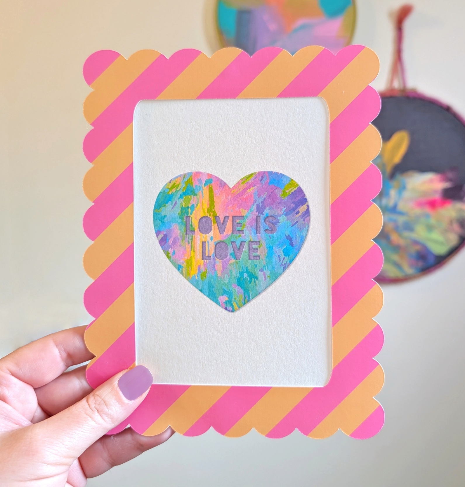 A close-up shot shows a hand holding a framed rainbow heart mini art print. The print features a colorful heart with "LOVE IS LOVE" text, central to the design. A pink and peach striped, scalloped frame surrounds the artwork. In the blurred background, hand-painted abstract circular wall art hangs on a white wall.