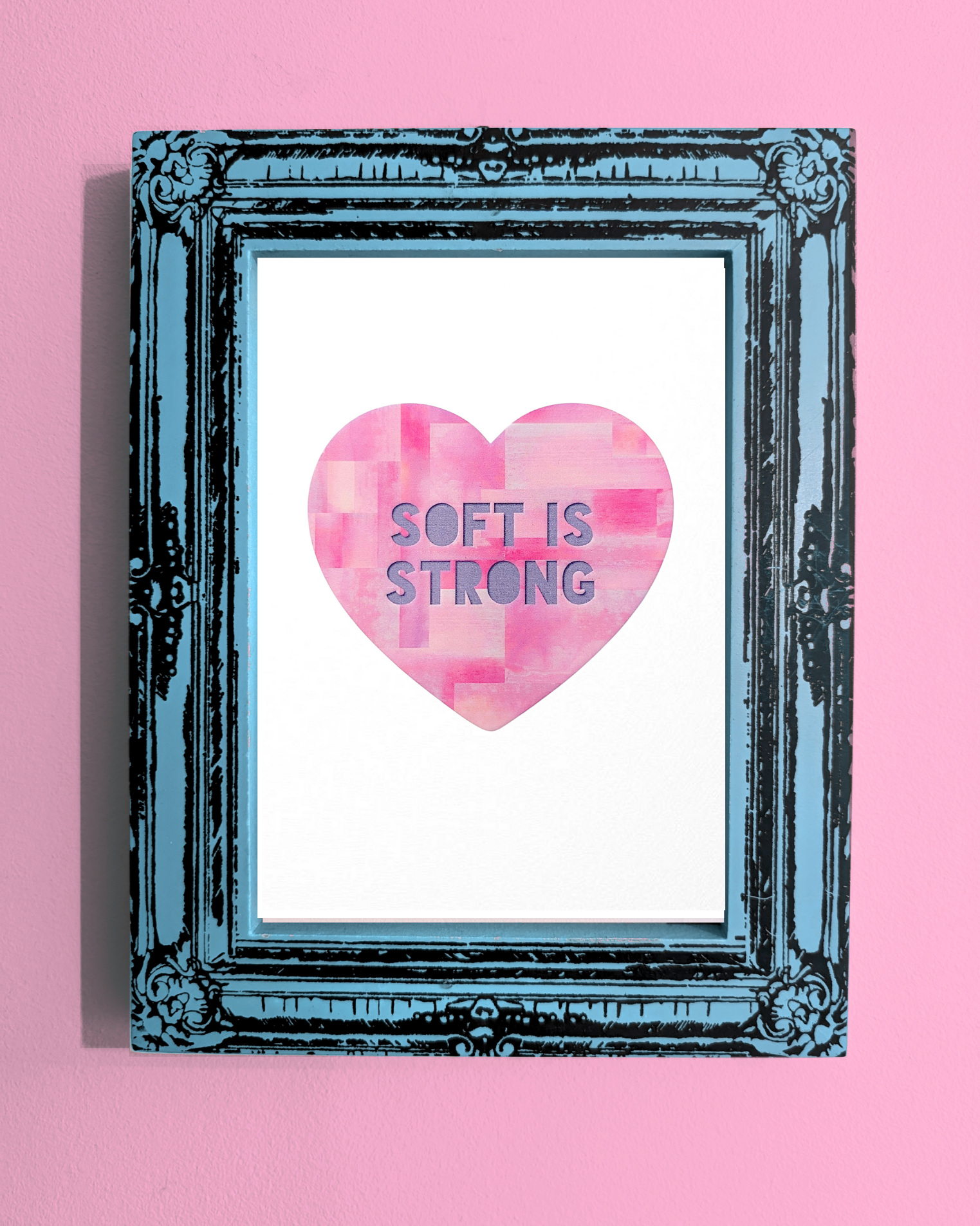 A framed art print displayed on a pink wall. The heart shape is filled with textured pink brush strokes and contains the phrase "SOFT IS STRONG". The ornate black frame has a distressed turquoise finish, adding contrast to the soft design.