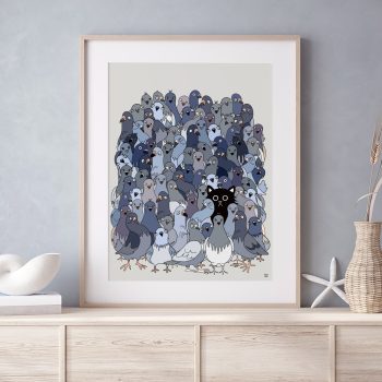 bf-cat-among-pigeons-scene-LIFESTYLE Framed illustrated art print of a large flock of grey and blue pigeons with a single black cat hidden among them, displayed in a light wood frame on a pale wooden sideboard with neutral home décor accessories.