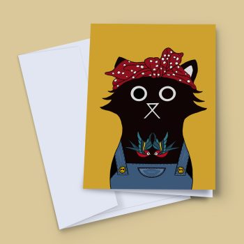 A greeting card featuring a bold illustrated black cat wearing blue denim dungarees and a red polka-dot headscarf, with a classic swallow tattoo on its chest, set against a mustard yellow background. The card is shown with a white envelope on a warm beige surface.