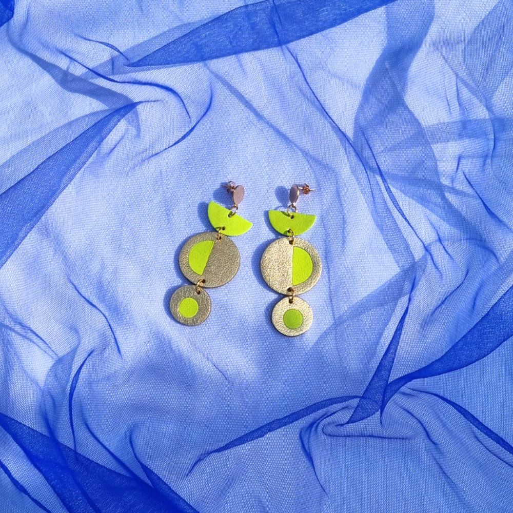 Geometric acid yellow and gold leather dangly earrings featuring a circular inlay design, resting on a draped bright blue tulle fabric background.