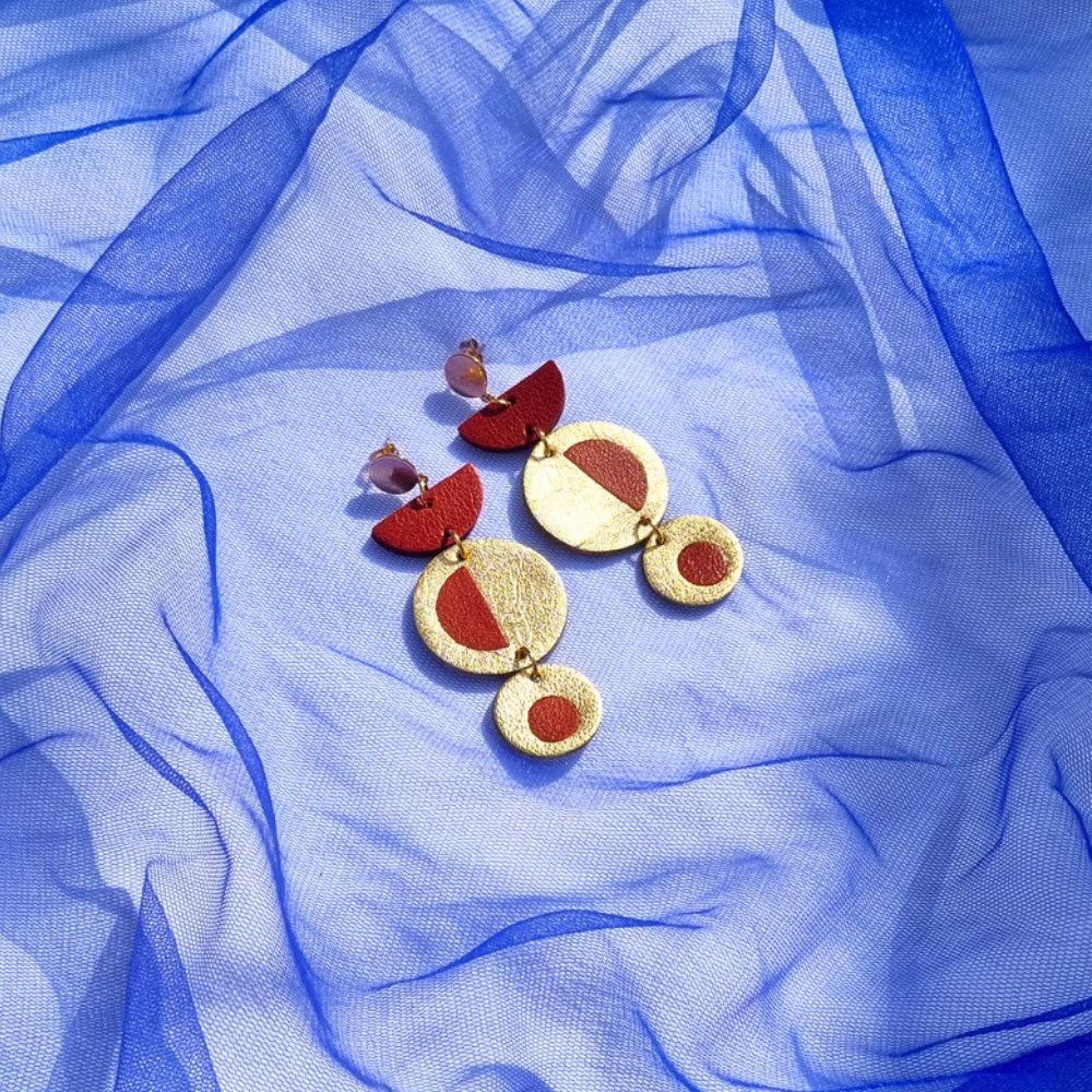 Handcrafted geometric dangly earrings with brick red inlayed in metallic gold leather, on a royal blue mesh fabric background.