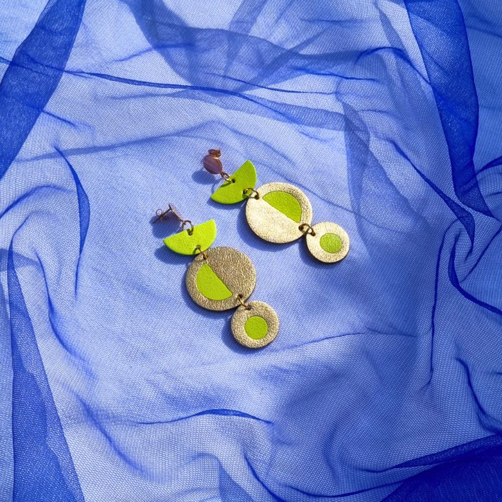 Geometric acid yellow and gold leather dangly earrings featuring a circular inlay design, resting on a draped bright blue tulle fabric background.
