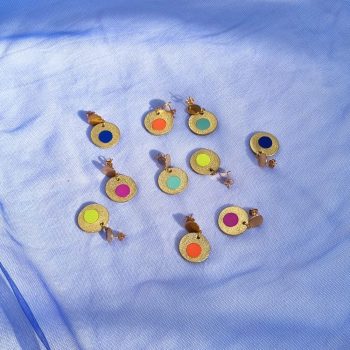 Five pairs of circular leather drop earrings in gold, featuring colourful centre inlays in orange, mint, navy, lime, and magenta, arranged on blue mesh fabric.